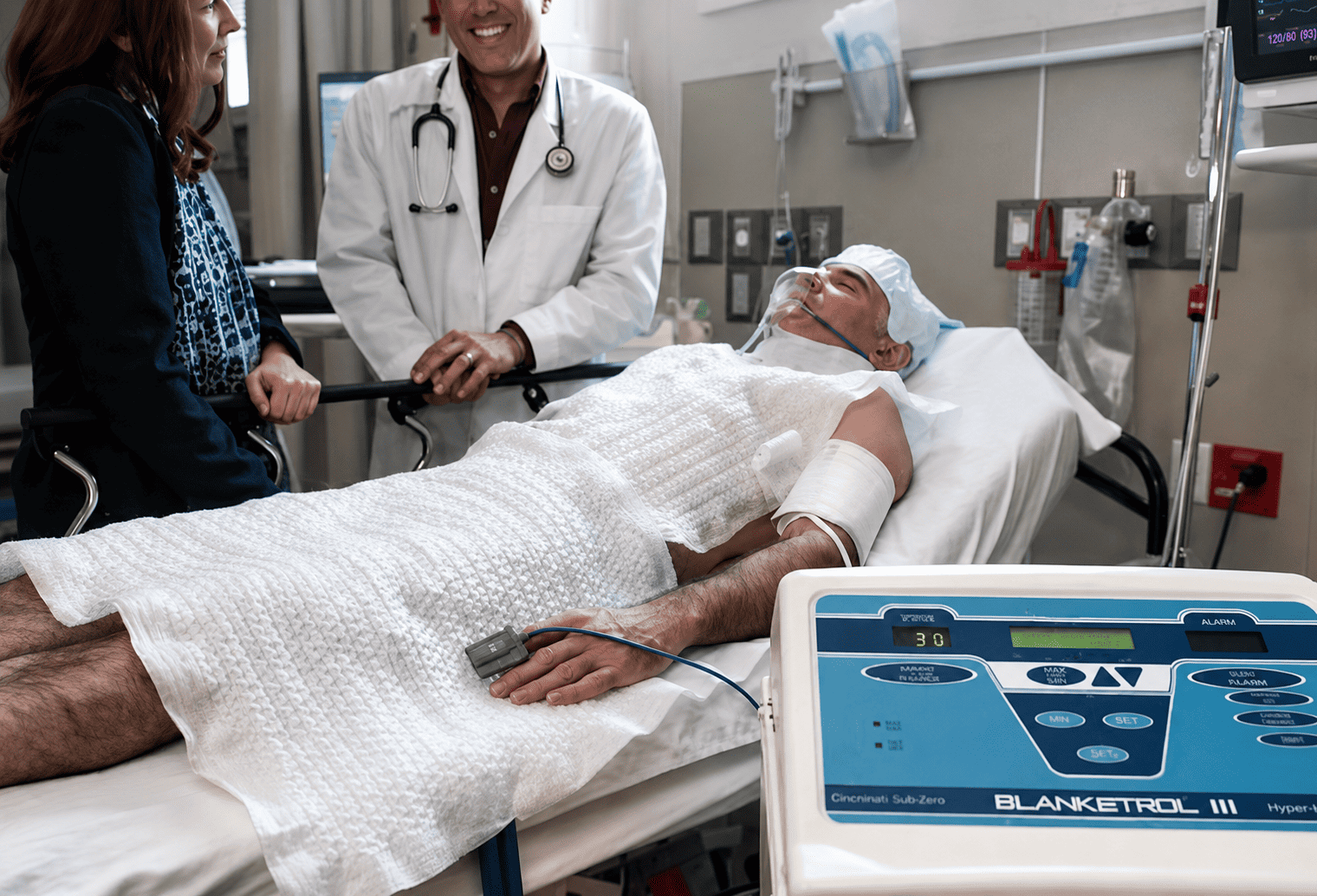 Medical team consulting with patient in hospital room with monitoring equipment.